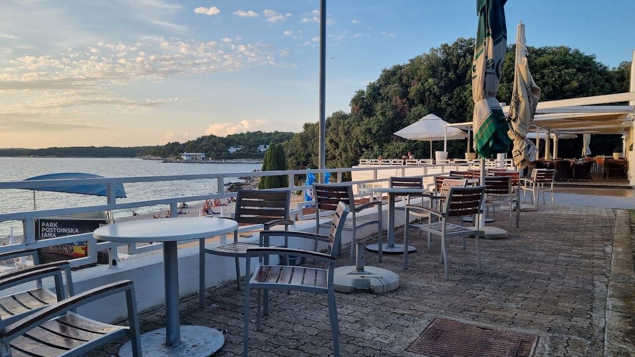 Tables and folded parasols on the Ambrela beach bar terrace above the pebble cove at Verudela in Pula