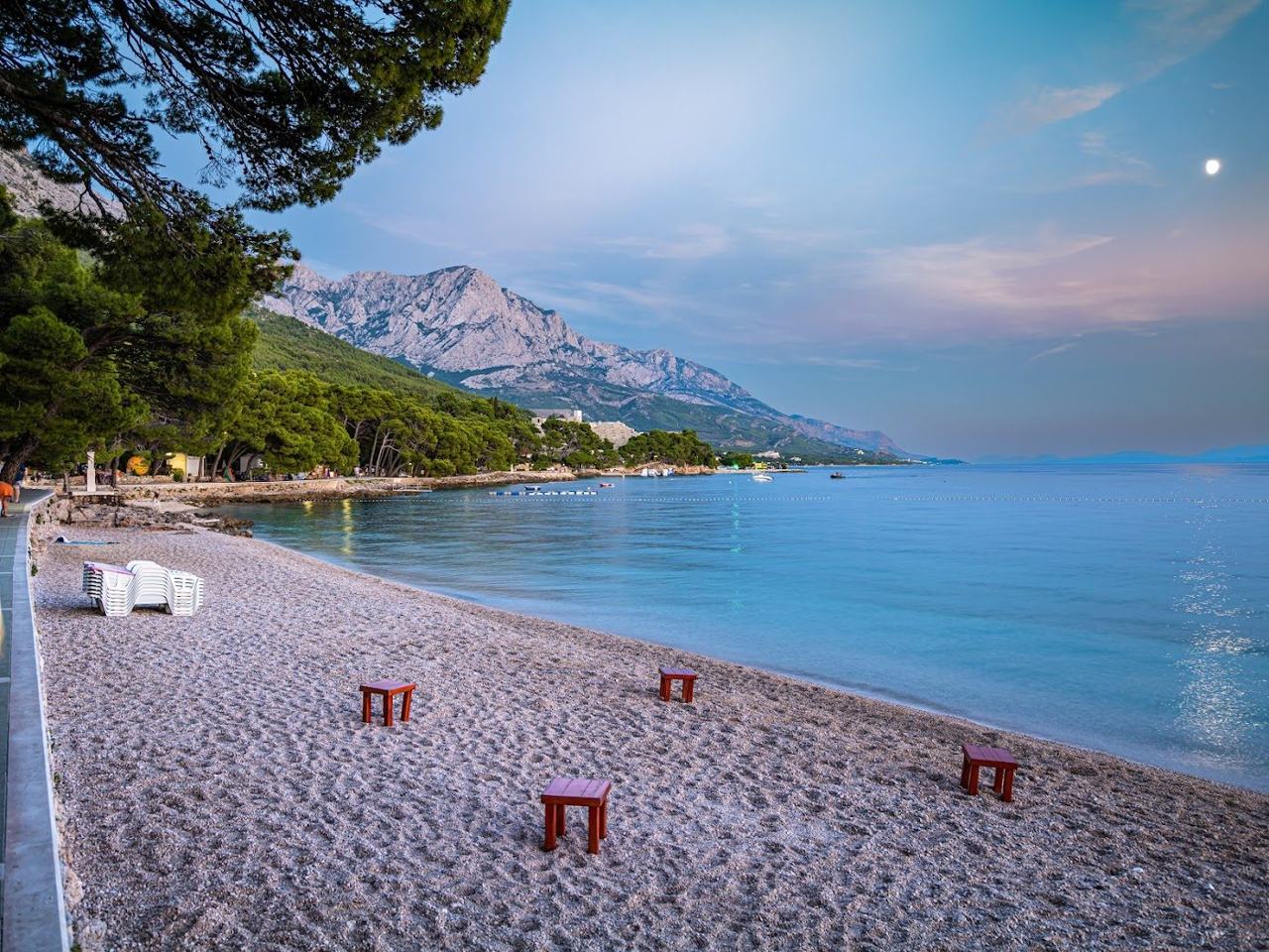 Punta Rata white pebble beach backed by pines with the pine topped Brela Stone in clear water at Brela Croatia